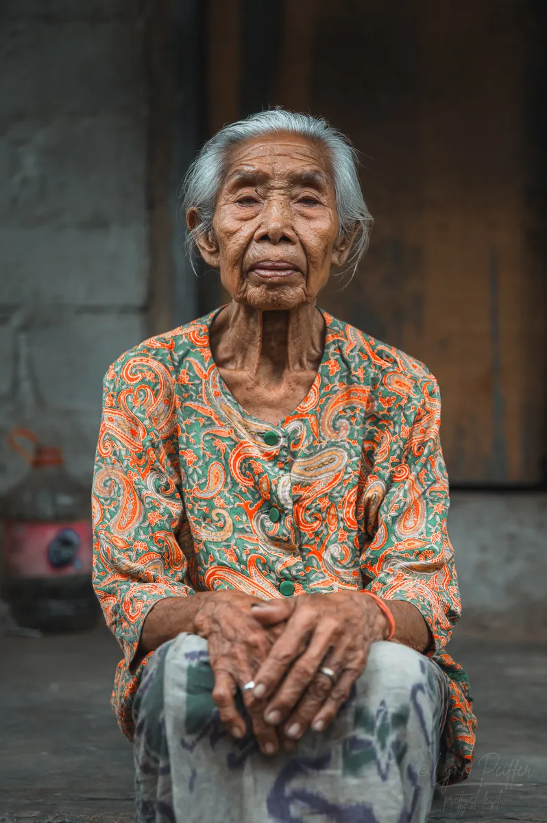 Portrait of an elderly Balinese woman with a weathered face wearing an orange and green paisley shirt and a sarong with her hands on her lap as she looks into the camera.
