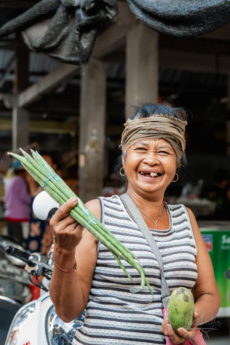 A Balinese woman smiles, displaying a few missing teeth, while holding up fresh produce at a market in Bali, Indonesia.