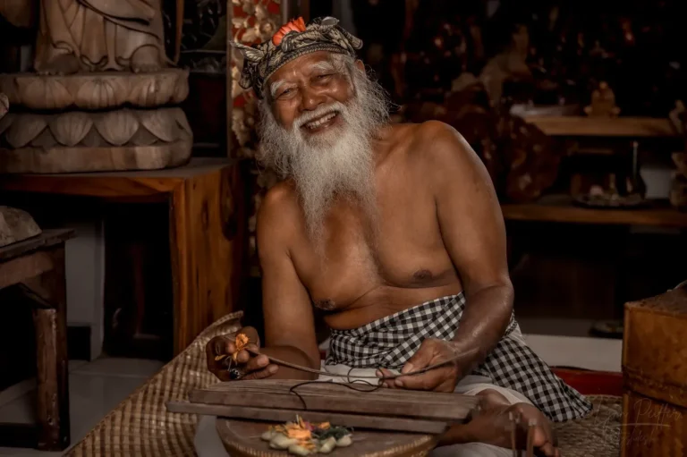 Portrait of a grinning elderly Balinese man wearing traditional ceremony attire as he sits in an artisan shop.