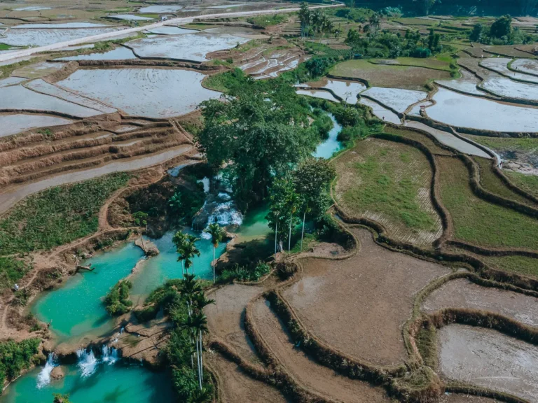 Intricate rice terraces surround a striking turquoise lagoon and waterfall in Sumba, Indonesia.