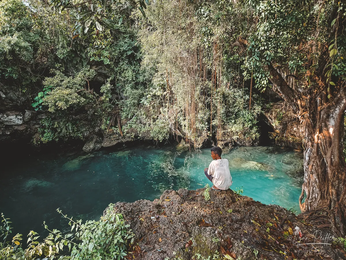 Sumba Indonesia Travel Photo 01 by Lynn Peiffer A Sumbanese man in a white t-shirt, grey shorts, and flip flops, crouches on the edge of an embankment overlooking a turquoise lagoon surrounded by tropical foliage.