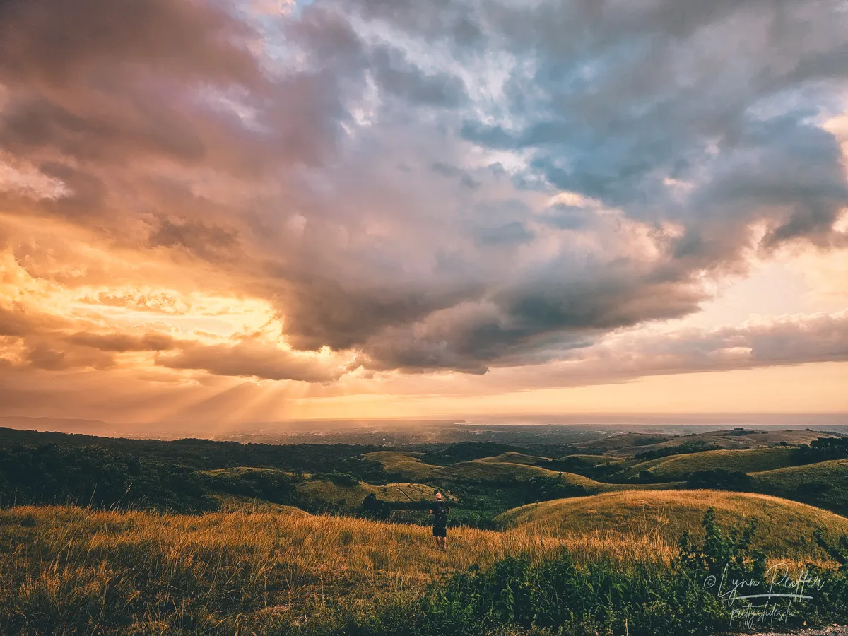 Sumba Indonesia Travel Photo 02 by Lynn Peiffer A landscape of the sunlight piercing clouds over rolling green hills with a man taking in the view in Sumba, Indonesia.