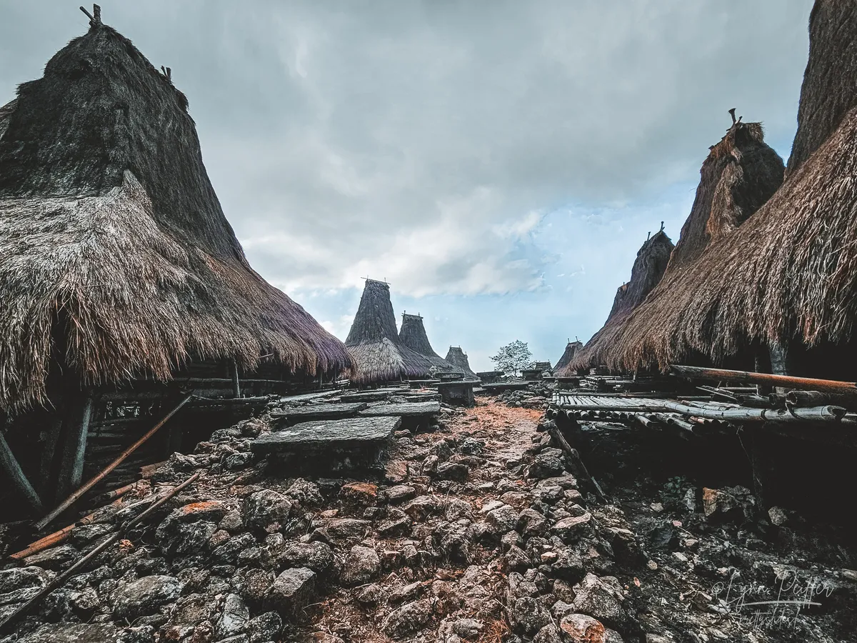 Sumba Indonesia Travel Photo 05 by Lynn Peiffer Stone rubble between two rows of high thatched alang-alang roofs of traditional Sumbanese houses against a cloudy sky.